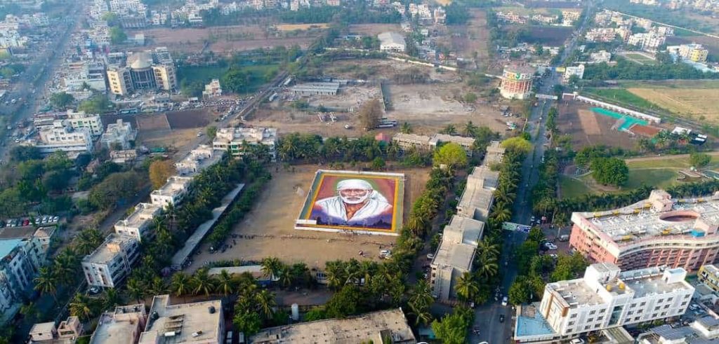 Sai baba images free download: Aerial drone view of massive ground rangoli portrait of Shirdi Sai Baba in white headcloth created with colored powders in large courtyard surrounded by buildings, trees, and sports field.