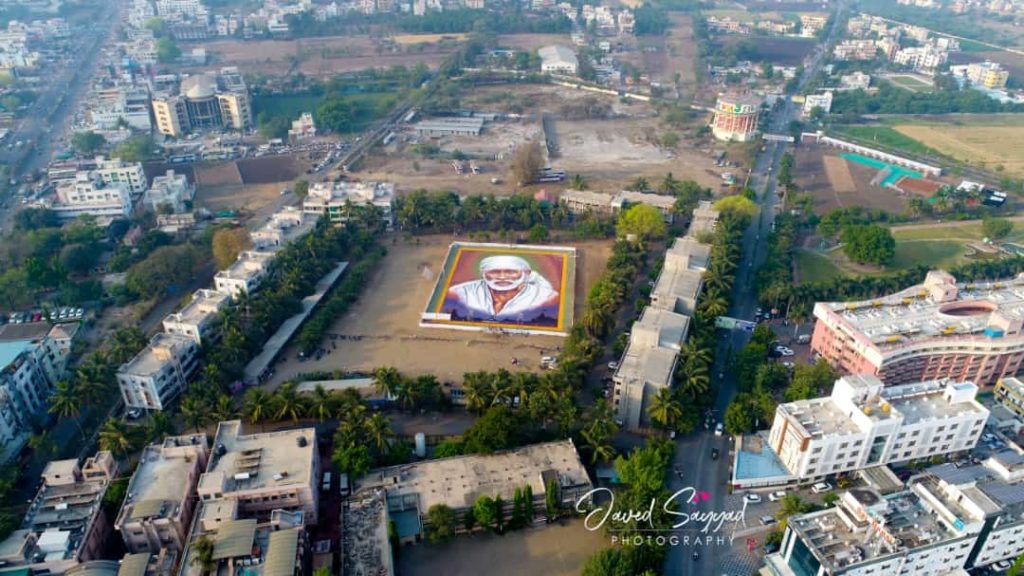 An aerial drone shot showing a giant, colorful portrait of Shirdi Sai Baba created as a rangoli in the center of a large open field. The massive artwork is surrounded by city buildings and trees, showcasing the scale of the devotional tribute.